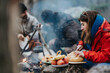 © qunica.com - A woman slices a loaf of bread beside a campfire with friends cooking in the background. The group prepares and shares an outdoor meal around a smoky fire.
