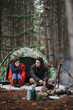 © qunica.com - A young couple sits at the entrance of a camouflage tent in a pine forest. The pair chat quietly beside a small campfire at their secluded campsite.