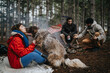 © qunica.com - A group of friends camping in a pine forest with a large dog interacting closely. Two people tend the campfire while a woman cuddles and kisses the dog beside a hammock.