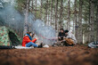 © qunica.com - A group of friends camping in a pine forest sits by a small campfire with a dog. Three people relax by a tent and hammock while preparing the fire in the woods.
