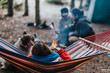 © qunica.com - A person relaxes in a colorful striped hammock while a brown dog sits nearby. Two friends chat by a small campfire in the background, creating a cozy outdoor scene.