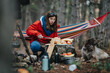 © qunica.com - A woman in a red jacket prepares food on a rock at a forest campsite while her dog watches. She chops bread and arranges fruit beside a hammock and a small campfire.