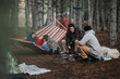 © qunica.com - Three friends relax around a campfire beside a colorful hammock with two dogs nearby. A casual pine forest camping scene showing conversation, warmth and outdoor leisure.