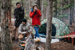 © qunica.com - Three friends camping in a forest beside a camouflaged tent and a small campfire. They prepare food and drink while standing and crouching near the fire during a chilly outdoor trip.