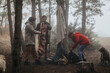 © qunica.com - A group of friends unpack and prepare camping gear in a foggy mountain forest clearing. They organize backpacks, a colorful rolled sleeping bag and supplies beside their dogs.