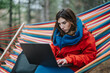 © qunica.com - A woman works intently on a laptop while sitting in a colorful hammock outdoors. She remotely works from a forest setting, bundled in a warm red jacket and scarf.