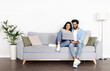 © Prostock-studio - Loving happy beautiful eastern couple in stylish casual outfit sitting on couch at home, embracing, using laptop, young indian man and woman watching movie online, copy space, full length