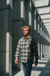 © f_bossa - Man wearing flannel shirt walks through concrete corridor while sunlight casts sharp shadows across the gray pillars. Golden hour brightness highlights his wavy hair.