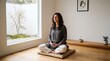 © Morepic - A serene woman practices meditation on a cushion in a minimalist room, featuring large windows overlooking a tranquil garden and incense burning nearby.