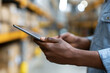 © lashkhidzetim - Hands hold tablet in warehouse setting. Fingers grip device with focused intent. Background shows stacked shelves and bright lights. Person appears engaged in work or inventory tasks