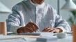 © CYBERPINK - Elderly man sitting at a desk and writing on a notebook. he is wearing a white shirt and has a beard. the desk is cluttered with papers, a lamp, and a plant.