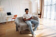 © standret - Young man with headphones sitting on a couch using smartphone in a modern living room with a desk and chair visible in the background