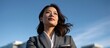 © avaye - Young businesswoman in gray blazer holding smartphone gazes confidently upwards outdoors against blue sky background