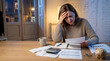 © itlada - Stressed woman calculating home finances at night, reviewing bills and expenses with a calculator, piggy bank, and coins on a wooden table, representing debt, inflation, and budget crisis