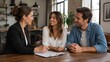 © olga_demina - A smiling woman in a black blazer signs a document on a clipboard while a young couple sits beside her at a wooden table inside a bright loft-style coffee shop, together happily