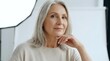 © Undersea - Mature woman with long gray hair and fair complexion, wearing casual beige top, hand near face in thoughtful pose, indoor setting with white background.