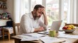 © Wankatsu - Stressed middle aged man looking at laptop and bills with a worried expression while sitting at a wooden table in a bright business lifestyle concept