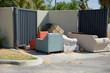 © Del Harper - Discarded sofas and armchairs sit beside a metal dumpster enclosure at a residential curb, surrounded by cardboard and trash, suggesting illegal dumping, neglect and bulky furniture disposal on a sunn