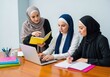 © yuwaree - Modest attire and religious headscarves are featured as a group works together using a laptop and colorful notebooks at a large wooden desk, classroom, desk, teamwork