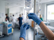 © Katerina - Healthcare worker adjusting an intravenous saline drip in a bright hospital ward. Close-up of gloved hands managing IV infusion equipment during patient treatment and clinical care.