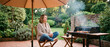 © itlada - Woman having cozy backyard barbecue under umbrella, sitting cross-legged on a wooden chair with a glass of red wine on a tiled patio, perfect for summer lifestyle and outdoor dining ads