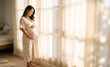 © Chari - Pregnant Asian woman in floral dress by sunlit window smiling