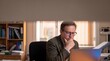 © benjamin - A thoughtful middle-aged man with glasses wearing a tweed jacket sits at his desk, intently focused on a laptop screen, with bookshelves filled with study materials in the background.