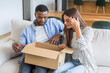 © simona - Young couple sitting on a sofa at home unpacking a cardboard box together, looking curious and excited while settling into their apartment