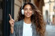 © btiger - young woman with long curly hair wearing white headphones and a gray plaid blazer points upward on a city street, confident and cheerful urban pose
