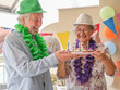 © luciano - Happy senior couple celebrating 75th birthday wearing festive hats and garlands, sharing moments of joy with a small cake and candles during an outdoor party