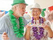 © luciano - Senior man with green hat and lei blowing out birthday candles for his wife's 75th birthday celebration, the happy woman smiling and wearing a sparkly hat and purple lei