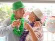 © luciano - Senior adult couple celebrating a special anniversary or birthday, embracing happily while toasting with champagne glasses and wearing festive party hats and leis outdoors