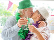 © luciano - Senior couple celebrating birthday, wearing festive hats and leis, sharing a romantic kiss while toasting with champagne, embracing lasting love and joy at a party