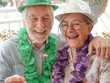 © luciano - Happy senior couple celebrating a birthday, wearing festive hats and leis, holding sparklers while enjoying a joyful party moment together outdoors