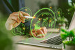 © Manasanan Wongchoomp - Business hand using stylus on laptop with green holographic CO2 dashboard, climate data, renewable energy icons, carbon reduction, sustainability technology and eco innovation concept.