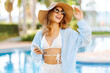 © maxbelchenko - Happy woman in hat and sunglasses enjoys lounging by pool, using her phone. Young woman in a hat and swimsuit sits by the pool with a smartphone. Concepts of relaxation, vacation, and luxury.