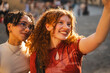 © Studio Marmellata - Two diverse young female friends smile warmly while taking a selfie together on a city street. The cheerful women enjoy a beautiful sunset during golden hour.
