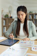 © Ratirat - Asian woman working intently on digital tablet using stylus pen at wooden desk with financial paper chart in comfortable modern home office environment