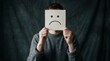 © Jafree - Man holding paper card with sad face emoticon covering his face. Depression, mental health, mood struggle and feeling unhappy concept. Expression of emotions and social anxiety mask