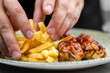 © pavel siamionov - Hands serving a plate of crispy golden french fries alongside grilled chicken pieces glazed in barbecue sauce.