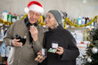 © JackF - Husband in a Santa hat and his wife choose perfume and a set of cosmetics in a supermarket against the background of a Christmas tree. Elderly couple choosing cosmetics for a gift for the New Year