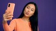© Yassine - Young asian woman holding a peach smartphone while pouting her lips for a playful selfie against a dark purple background