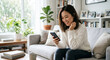 © Akash Tholiya - Smiling Young Asian Woman in White Knit Sweater Holding Smartphone in Bright Living Room, Casual Lifestyle Portrait, Natural Light