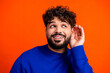 © deagreez - Young man listening intently with hand to ear in bright portrait, curious expression, blue sweater against orange background conveying communication and attentive listening