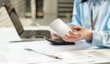 © Father_Studio - Businesswoman hands review receipts beside laptop and calculator. Focused expression shows careful financial management. Desk cluttered with papers implies busy work environment.