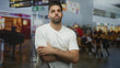 © Krakenimages.com - Man with arms crossed standing in airport departure lounge wearing white t shirt and beard, looking at camera; confidence resilience.