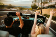 © Natalie Wheeler - Young couple driving in convertible with hands in the air