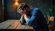 © miss irine - Young man sits at wooden table holding head in hands feeling overwhelmed. Stressed person suffers from depression in dark room. Upset male experiences anxiety frustration, pain. Lonely guy shows
