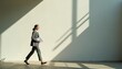 © miss irine - Businesswoman walks along white wall holding white documents. Female manager in grey formal suit strides across office floor during workday. Successful career woman moves forward in sunlight with