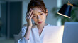 © k-studio - Exhausted Japanese woman touching her forehead while suffering from a headache at night office